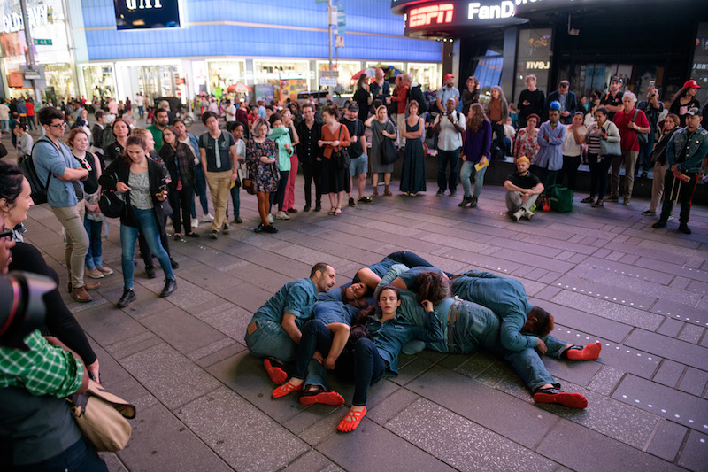 A group of dancers lay on the ground. They wear red shoes and jeans and jean button ups.
